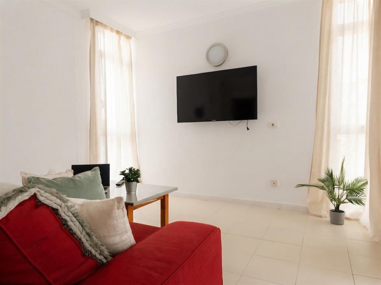 Ein Wohnzimmer mit einem roten Sofa, einem kleinen Tisch, einer Zimmerpflanze und einem Wandfernseher.