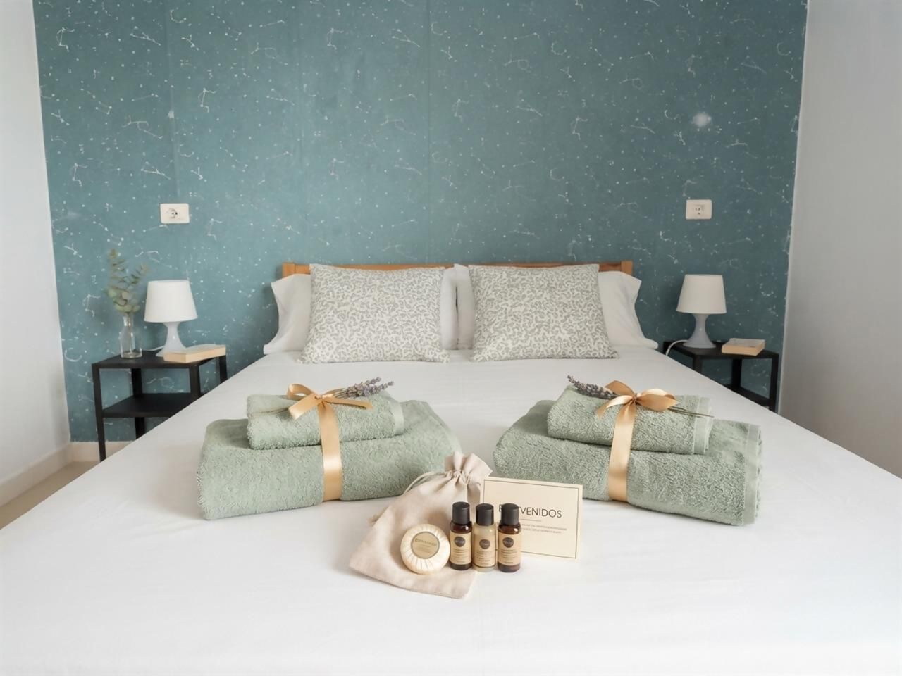 A neatly made bed with two pillows, a bedside table with a lamp, and a gift basket with toiletries on a hotel room.