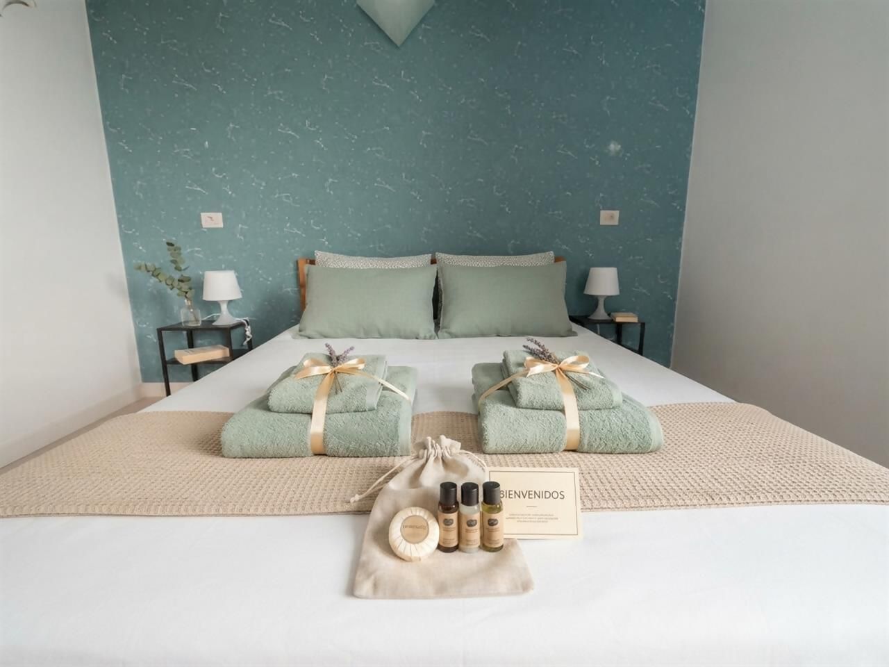 A neatly made bed with green towels, a welcome bag, and a sign.