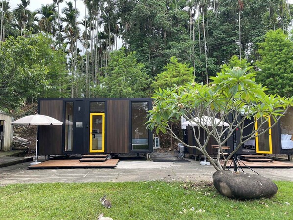Een moderne, compacte hut met een donkere buitenkant en gele accenten, omgeven door weelderig groen en hoge bomen.