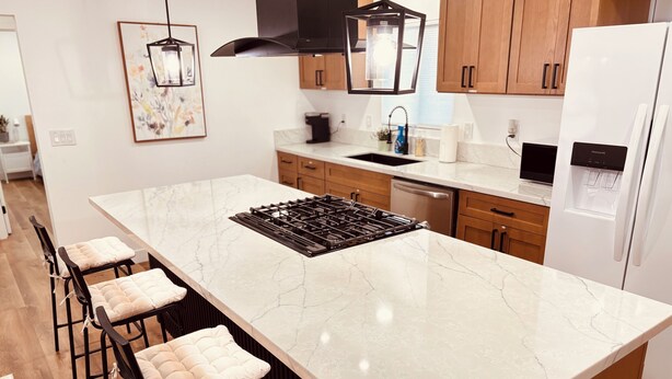 A modern kitchen with a large island, bar stools, wooden cabinets, a built-in stove, and pendant lighting.