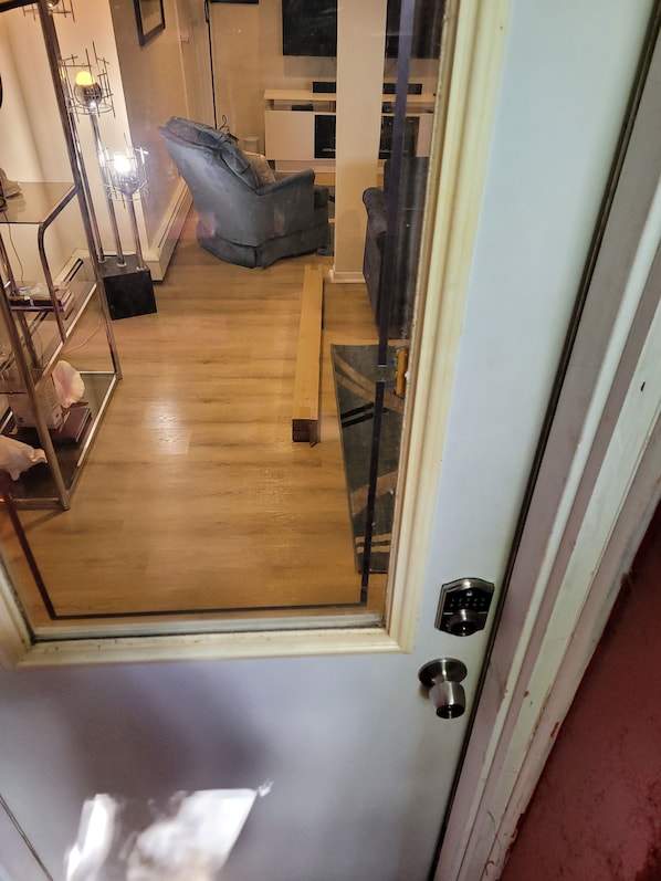 Vista a través de una puerta de cristal hacia una habitación con suelo de madera, un sofá gris, una mesita pequeña y un mueble blanco.