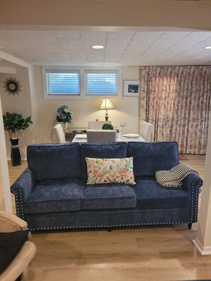 Una sala de estar con un sofá azul, una mesa de comedor con sillas, una lámpara y una cortina con estampado floral.