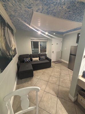 Un salon avec un canapé, un fauteuil, une fenêtre, une porte et un réfrigérateur.