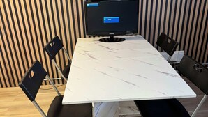 Un salón de conferencias con una mesa blanca, sillas negras, un monitor Samsung y una pared de madera a rayas.