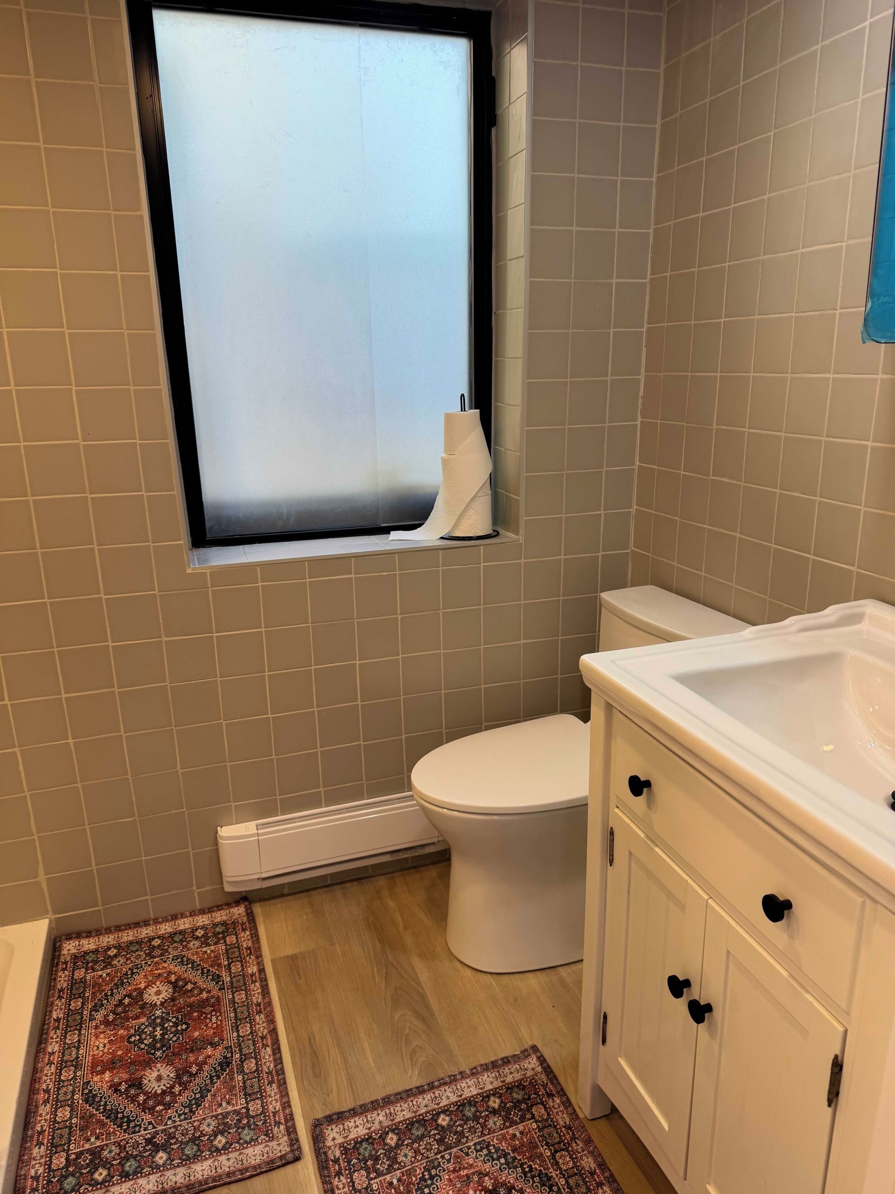 A bathroom with a toilet, a sink, a window with frosted glass, and two rugs on the floor.