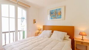 A bedroom with a large bed, a nightstand with a lamp, a window with a view, and a painting on the wall. - 2-room apartment with sea view - A/C, Loggia (PORT GRIMAUD)