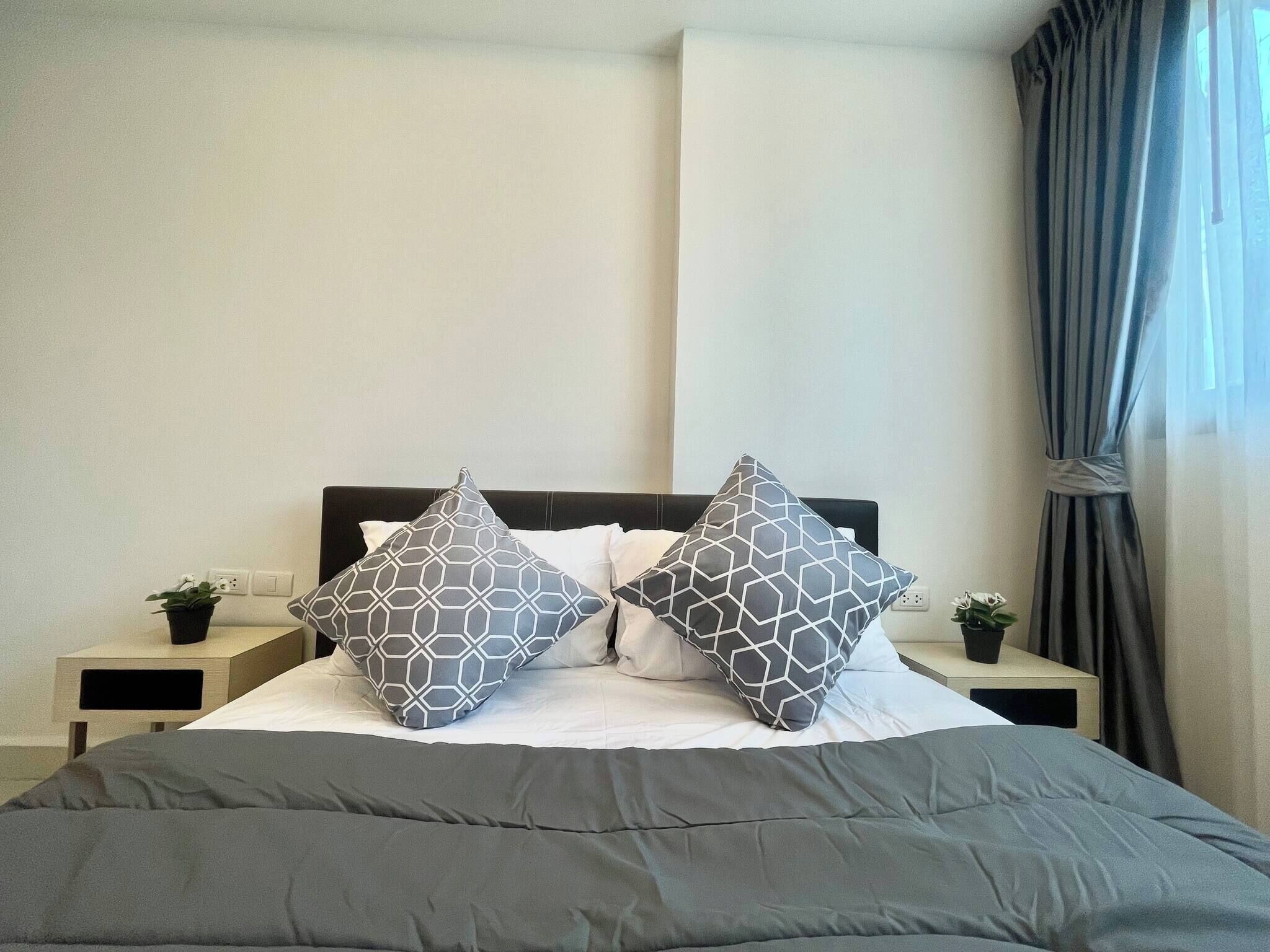 A neatly made bed with a grey comforter and two patterned pillows, a bedside table with a plant, and a window with curtains.