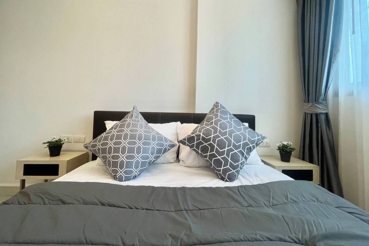 A neatly made bed with a grey comforter and two patterned pillows, a bedside table with a plant, and a window with curtains.
