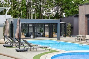 Un espace piscine moderne avec des chaises longues, un coin salon abrité et une vue sur les arbres environnants.