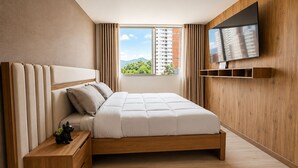 Ein modernes Schlafzimmer mit einem Holzkopfteil, einem Bett mit weißer Bettwäsche, einem Nachttisch, einem an der Wand montierten Fernseher und einem Fenster mit Blick auf Gebäude und Grünflächen.