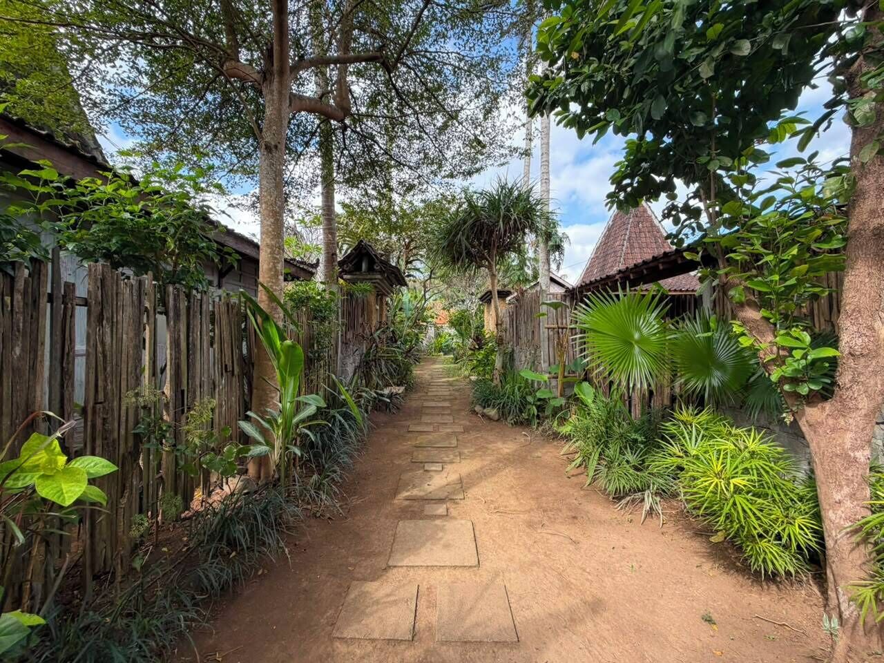 A narrow dirt path flanked by wooden fences and lush greenery, leading to traditional huts with thatched roofs.