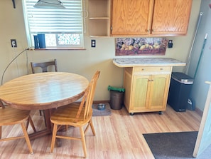 Un coin repas avec une table ronde en bois et des chaises, un buffet en bois orné d’une peinture représentant des fruits, et une fenêtre avec des stores.