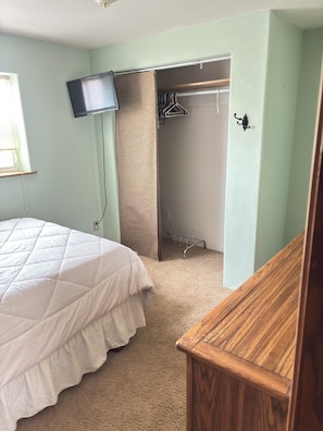 A bedroom with a bed, a TV mounted on the wall, a wooden dresser, and a closet with a clothes rack.