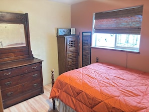 Une chambre à coucher avec une commode en bois, un lit recouvert d’une courtepointe orange, une fenêtre avec des stores et une armoire en bois.