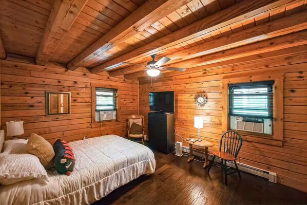 A wooden cabin bedroom with a bed, a chair, a table, a ceiling fan, and two windows with blinds.