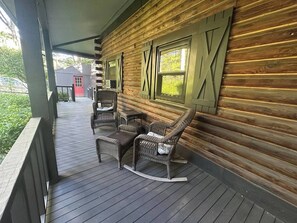 Eine Holzhütte mit Veranda, ausgestattet mit Korbmöbeln und Blick auf ein rotes Gebäude in der Ferne.