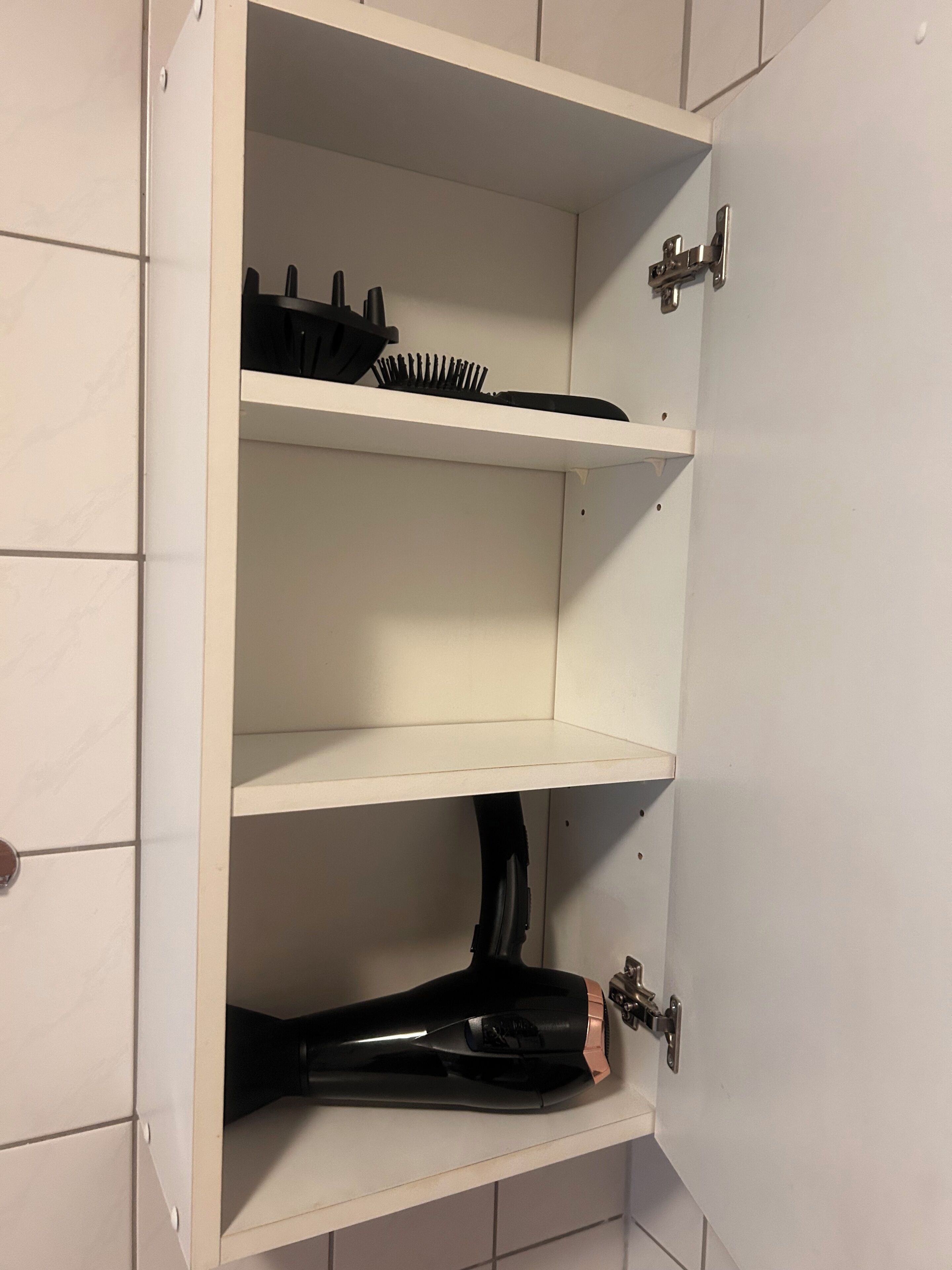 Een witte kast met drie planken, waarvan er één een zwarte haardroger en een zwarte haarborstel bevat.