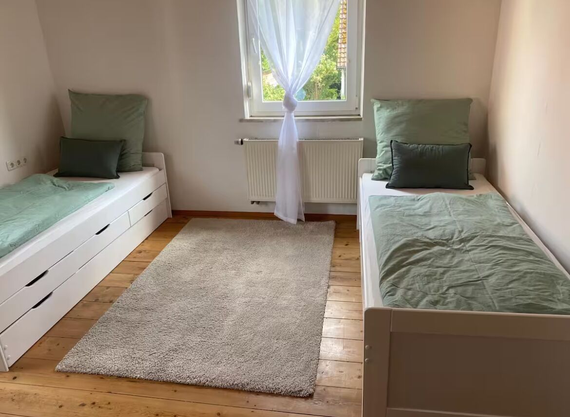 Ein Schlafzimmer mit zwei Einzelbetten, einem Fenster mit durchsichtigen Vorhängen, einem Heizkörper und einem beigen Teppich.