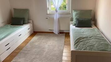 Ein Schlafzimmer mit zwei Einzelbetten, einem Fenster mit durchsichtigen Vorhängen, einem Heizkörper und einem beigen Teppich.