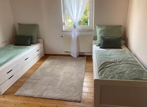 Ein Schlafzimmer mit zwei Einzelbetten, einem Fenster mit durchsichtigen Vorhängen, einem Heizkörper und einem beigen Teppich.