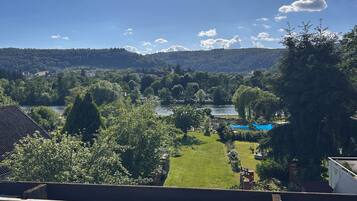 Blick von einem Balkon auf eine grüne Landschaft mit Bäumen, einem Fluss und fernen Hügeln.