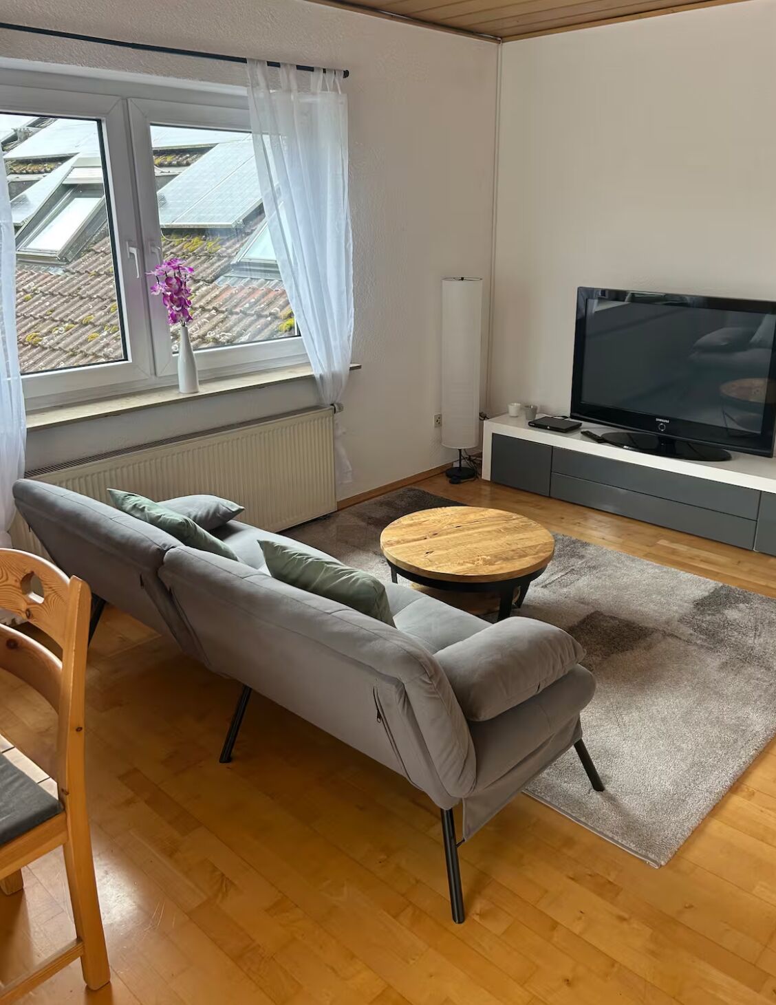Ein modernes Wohnzimmer mit einem grauen Sofa, einem runden Couchtisch, einem Flachbildfernseher und Blick auf ein Dach durch ein Fenster.