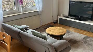Ein modernes Wohnzimmer mit einem grauen Sofa, einem runden Couchtisch, einem Flachbildfernseher und Blick auf ein Dach durch ein Fenster.