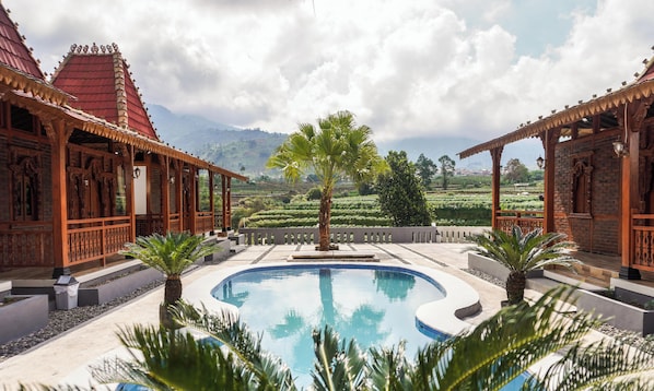 A pool with a view of terraced fields, wooden buildings with intricate designs, and a palm tree in the center. - Instay Villa Dieng Kademangan (Wonosobo)