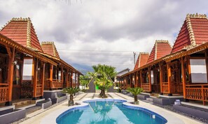 Uma piscina com dois edifícios de madeira idênticos em ambos os lados, céu claro e uma palmeira no centro.