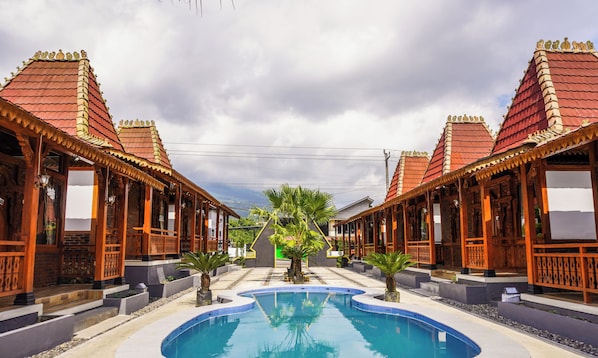 A pool with two identical wooden buildings on either side, a clear sky, and a palm tree in the center. - Instay Villa Dieng Kademangan (Wonosobo)