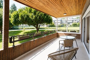 Een overdekt buitengebied met een tafel en stoelen, uitzicht op een groen park en een bakstenen muur.