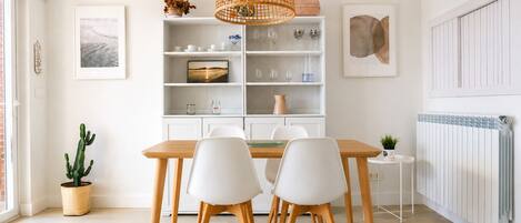 Un comedor con mesa de madera y sillas blancas, un estante blanco con cristalería, una lámpara colgante tejida y una planta de cactus.