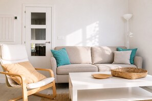 Een woonkamer met een beige bank, een houten stoel, een witte salontafel en een gevlochten mand.