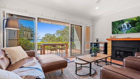 A modern living room with a sofa, coffee table, and fireplace. There is a sliding glass door leading to an outdoor area with a table and chairs.