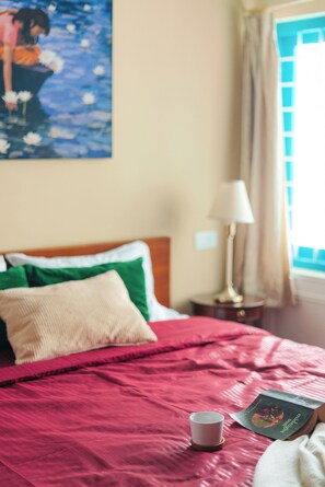 A bed with a red bedspread, a white pillow, and a green pillow. There is a book and a cup on the bed.