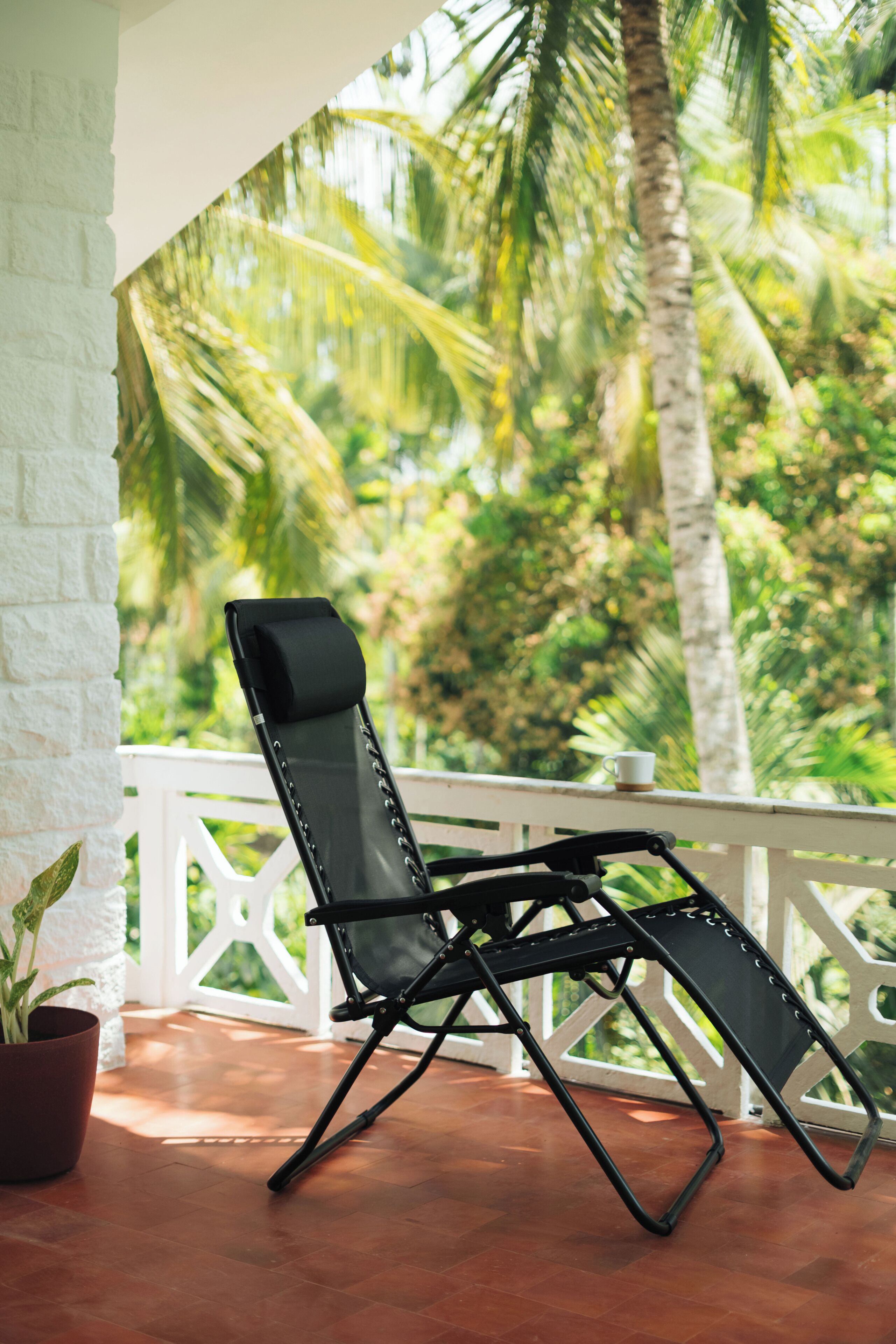A black lounge chair on a wooden deck with a white railing, overlooking a lush green landscape with palm trees.