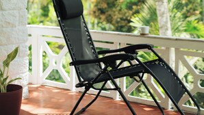 Una silla de lounge negra sobre una terraza de madera con barandilla blanca, con vistas a un exuberante paisaje verde con palmeras.