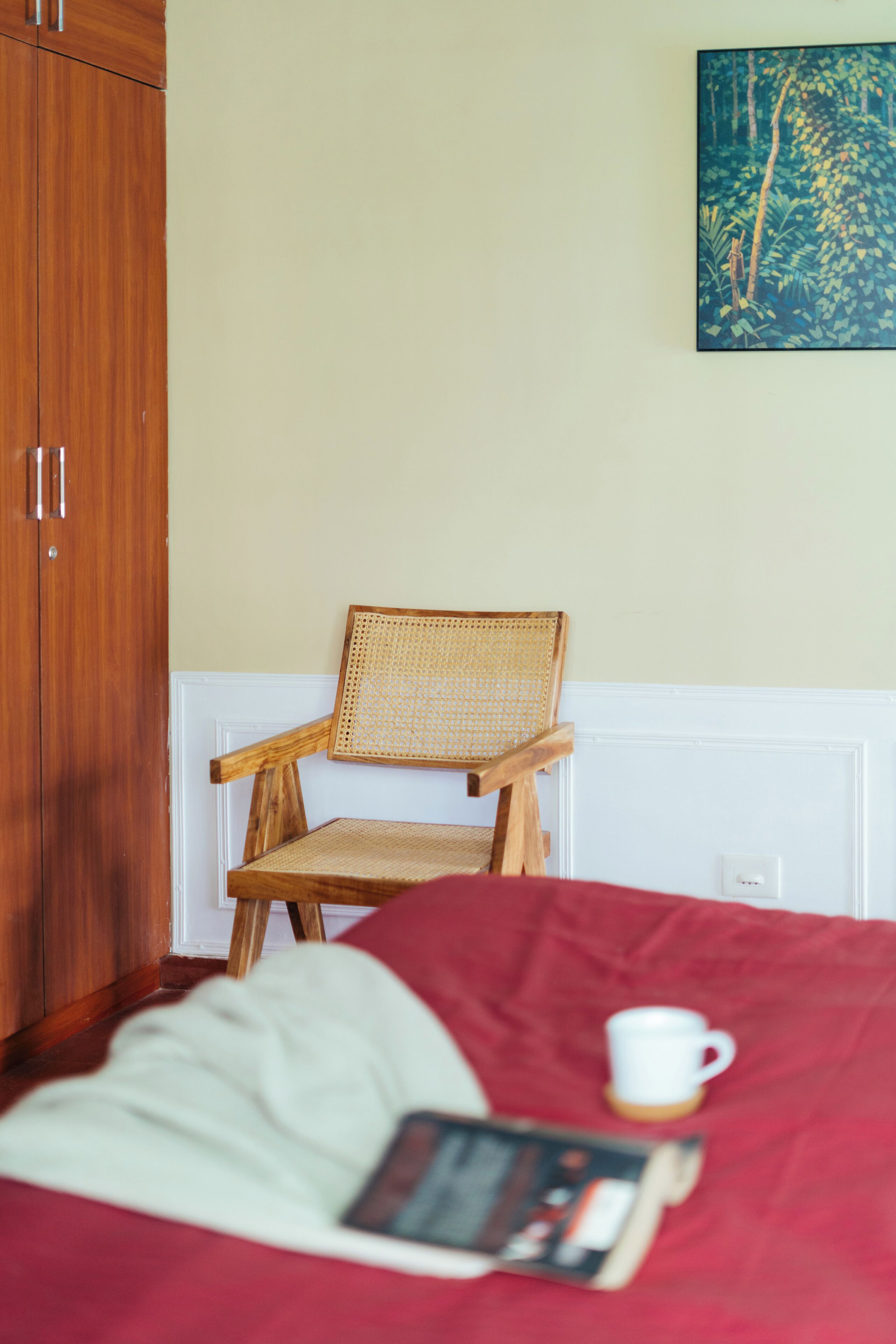 Una habitación con una silla de madera, una colcha roja, una pared blanca y un cuadro.