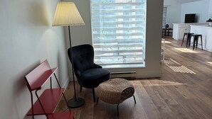 A modern living room with a black chair, a red shelf, a wicker ottoman, and a large window with blinds.