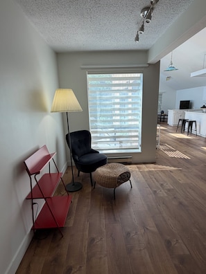 A modern living room with a black chair, a red shelf, a wicker ottoman, and a large window with blinds.