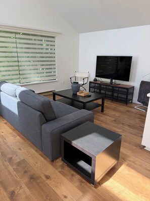 Un salon moderne avec un canapé, une table basse, un téléviseur et une fenêtre équipée de stores.