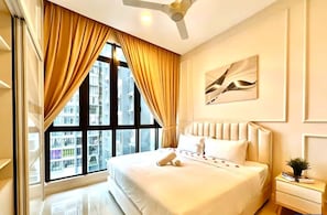 A hotel room with a large bed, a ceiling fan, a window with curtains, a nightstand with a book, and a wall-mounted artwork.
