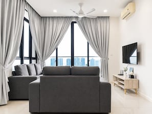 A modern living room with a grey sofa, large windows with curtains, a ceiling fan, a flat-screen TV, and a small wooden side table.