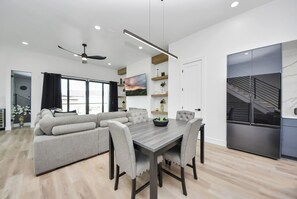 Una cocina moderna con refrigerador empotrado, un comedor con mesa y sillas de madera, y una sala de estar con sofá y ventilador de techo.