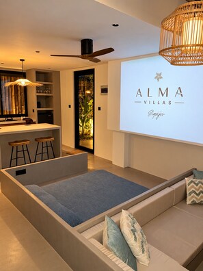 Una sala moderna con sofá, una mesa de centro y una zona de bar con taburetes. Hay un ventilador de techo y una pantalla de proyector mostrando 'ALMA VILLAS'.