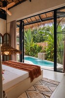 Un dormitorio con una cama grande, vista a una piscina y un jardín tropical afuera.