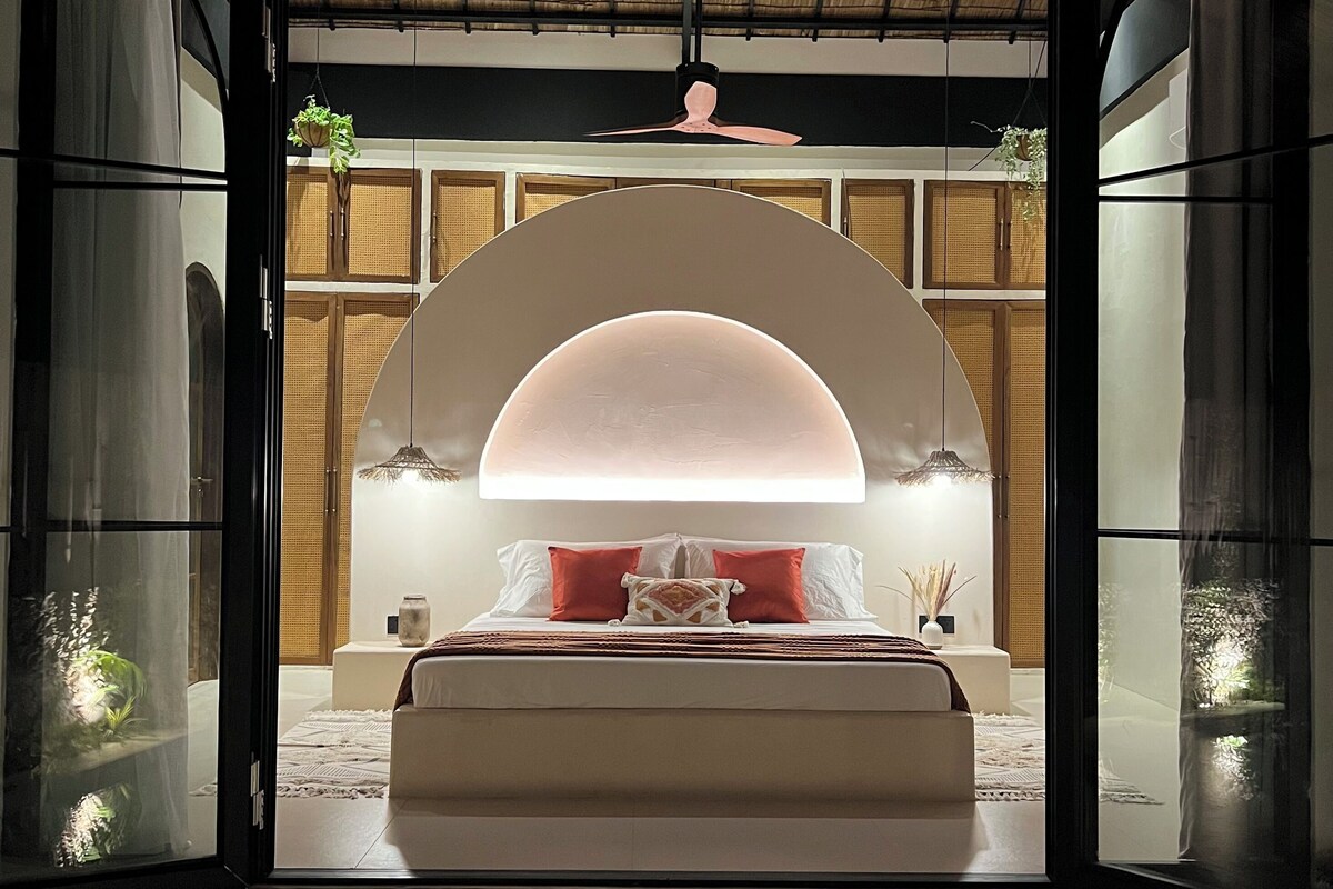 A modern bedroom with a bed centered under a large arched window, a ceiling fan, and a view of a traditional room with a thatched roof.