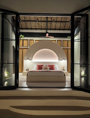 A modern bedroom with a bed centered under a large arched window, a ceiling fan, and a view of a traditional room with a thatched roof. - Alma Villas (San Juan)