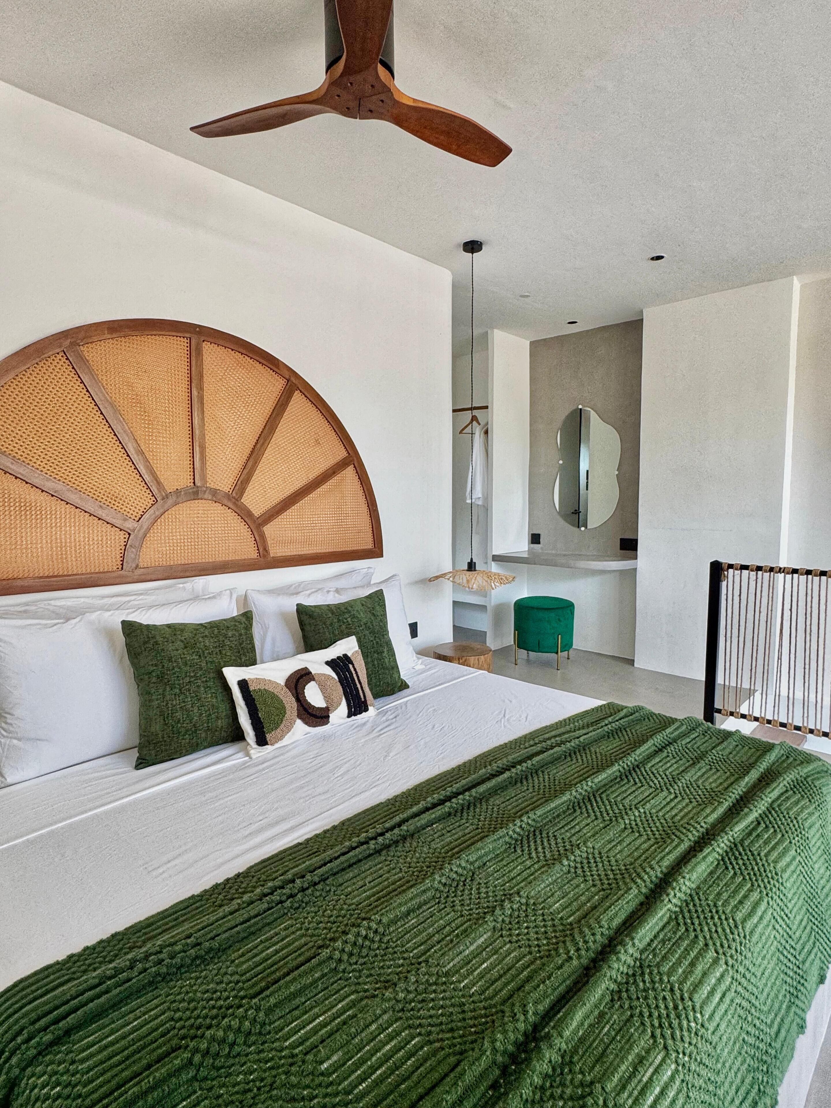 A modern bedroom with a large bed, a wooden ceiling fan, a round woven headboard, and a green throw blanket.